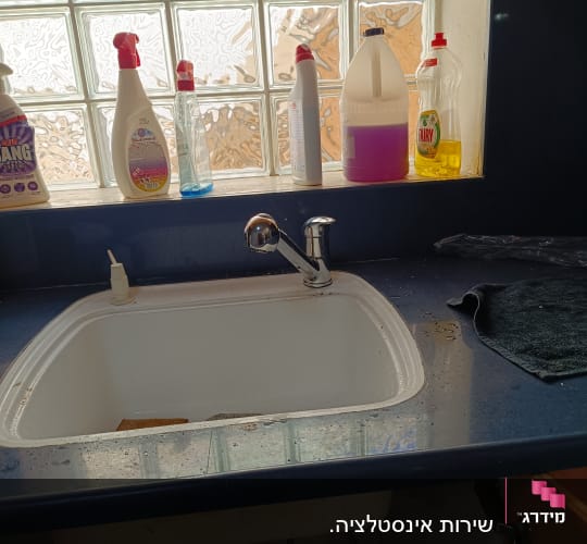 כיור מטבח עם ברז וחומרי ניקוי על אדן החלון